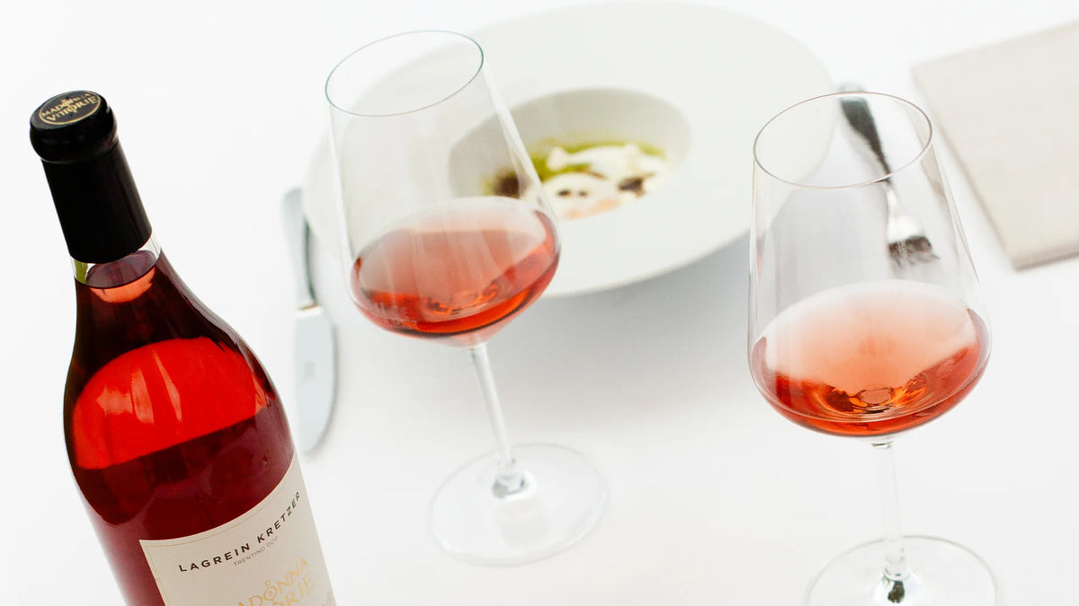 Bottiglia di vino rosato MDV accanto a due calici colmi su tavolo bianco con piatto sullo sfondo